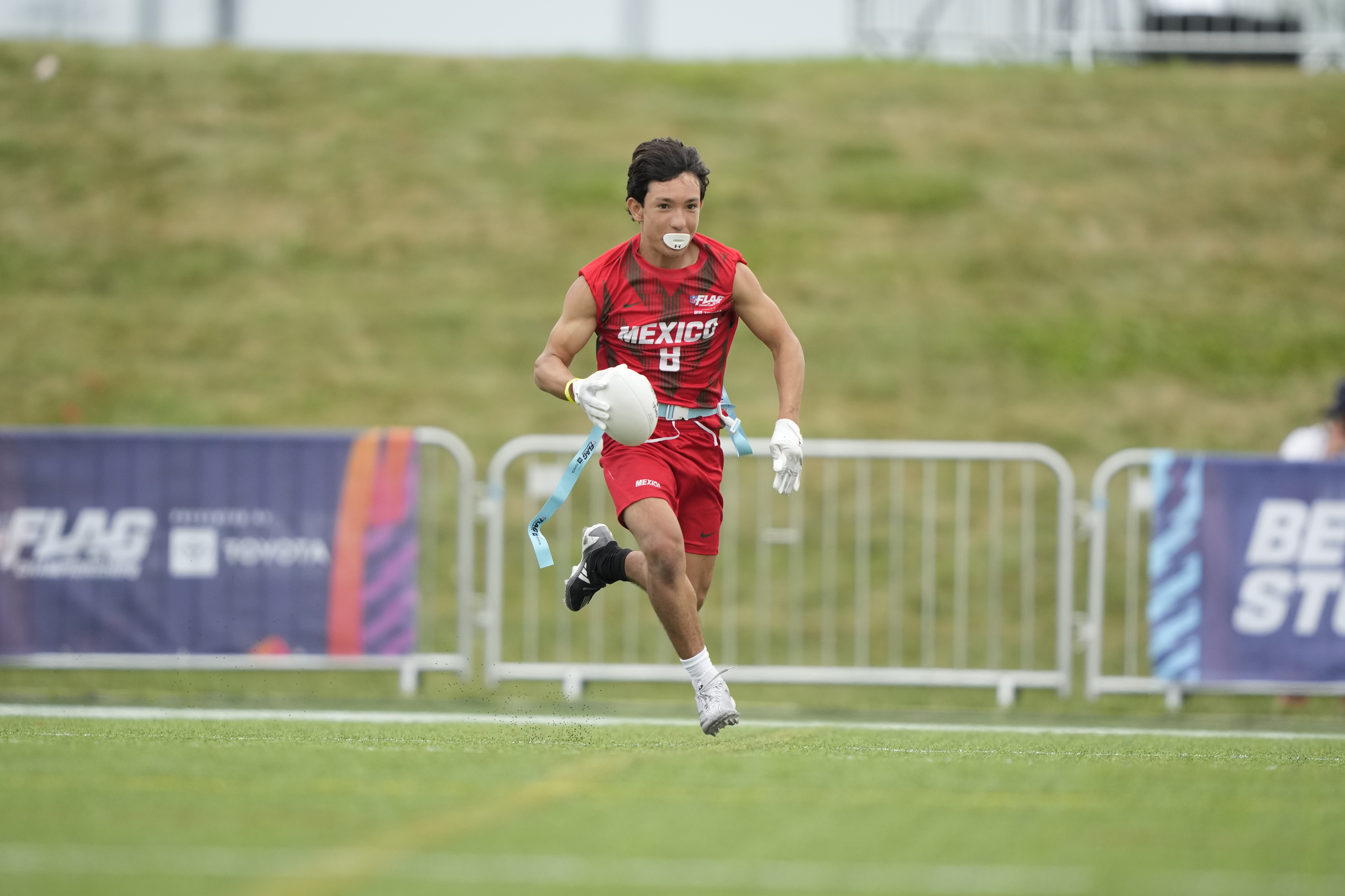 Jugador del equipo México con el dorsal 8 corre a toda velocidad durante el Campeonato Mundial de NFL FLAG, con sus banderas colgando del cinturón y el logotipo del torneo visible al fondo.