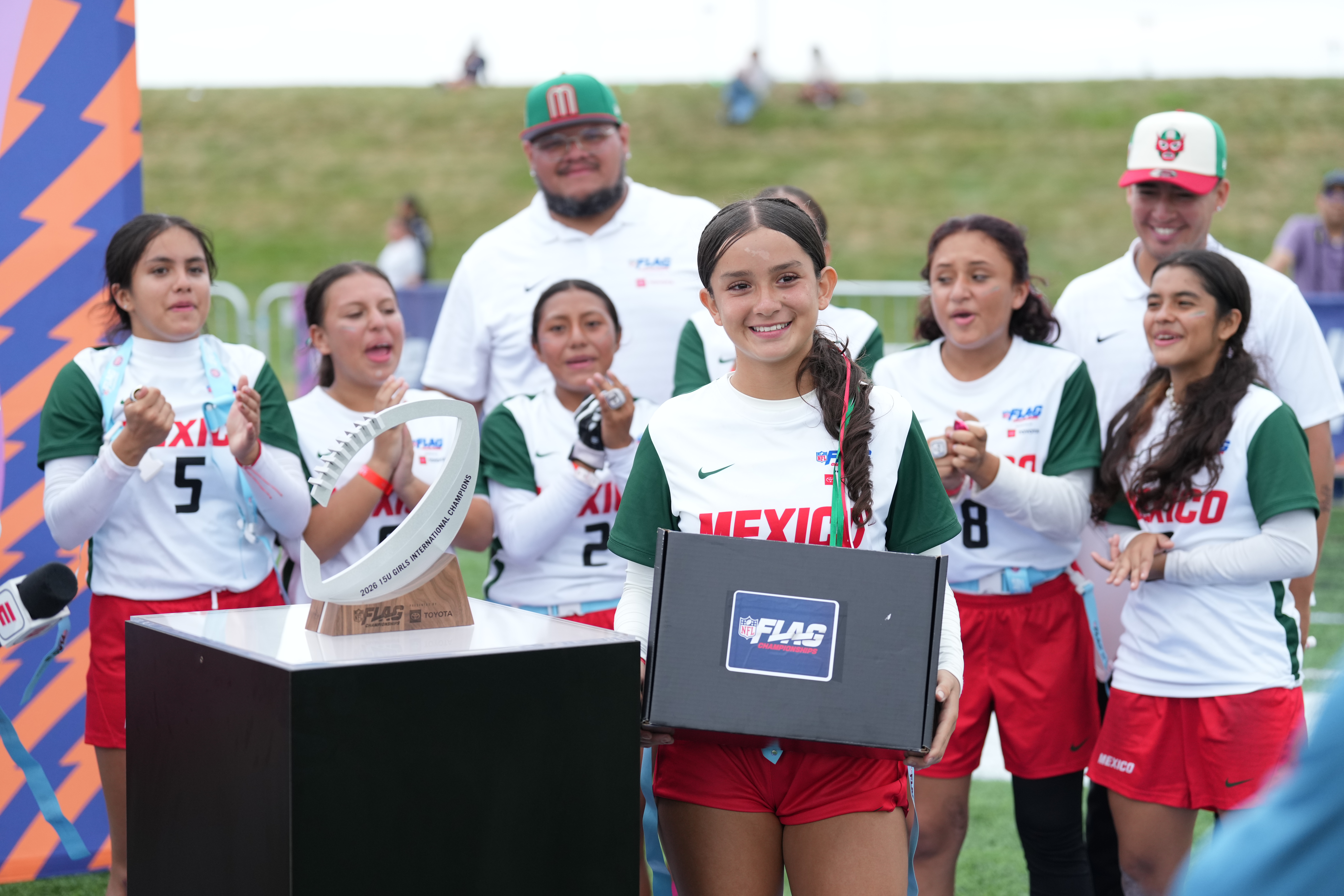 Jugadora del equipo México sonríe frente al trofeo de Campeonas Internacionales Sub-15 Femenil 2026 de NFL FLAG, mientras sus compañeras y cuerpo técnico celebran detrás de ella.
